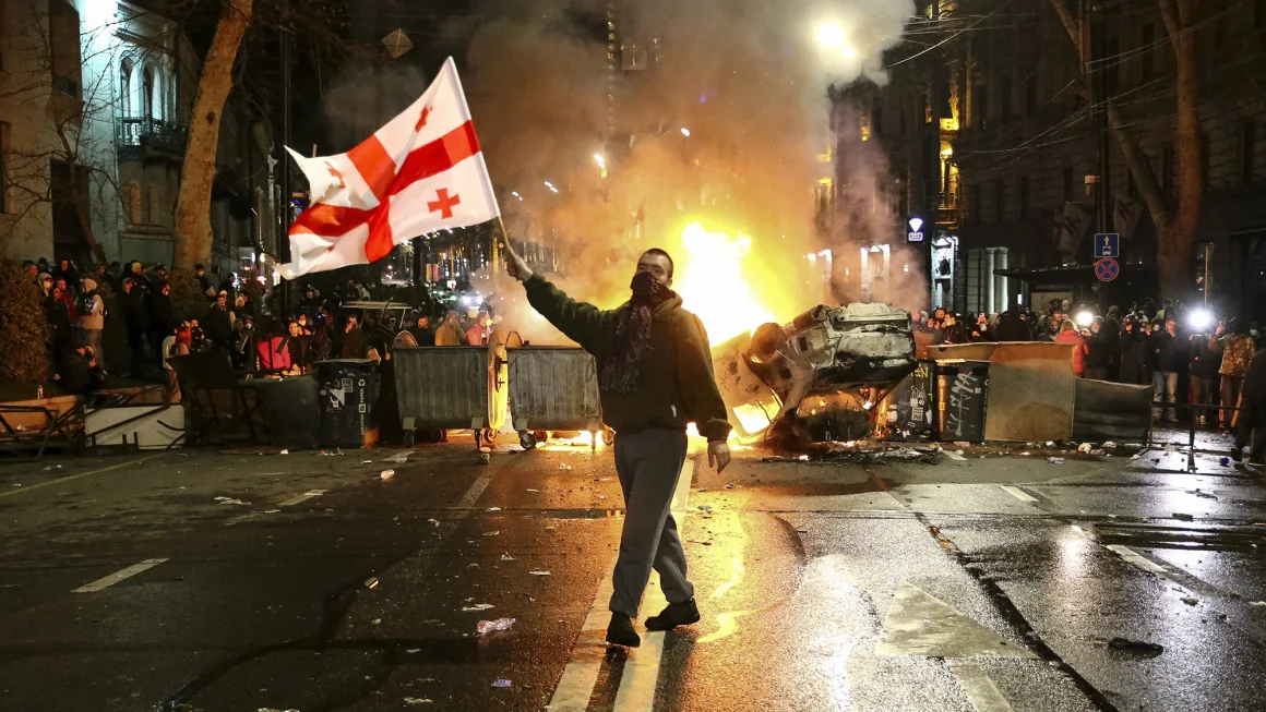 Georgian National Flag Waves In Front Of a Burning Barricade Not Far Away From The Parliament Building In Tbilisi